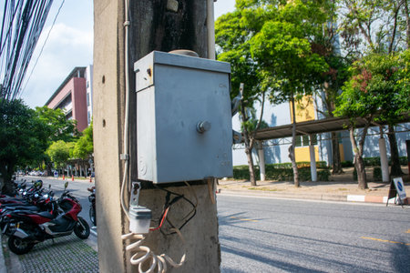 Image of a gray electrical box mounted on a utility pole in an urban setting, surrounded by green trees and parked motorcycles, showcasing city life.の写真素材