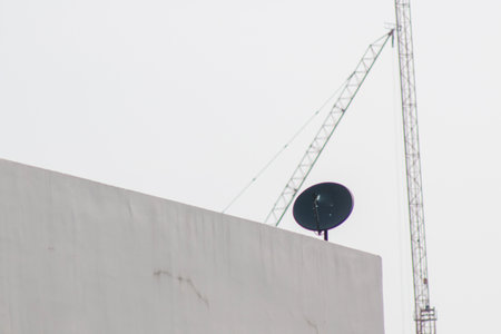 A satellite dish positioned on a rooftop with a construction crane visible in the background, symbolizing urban development and technological connectivity in modern architecture.の写真素材
