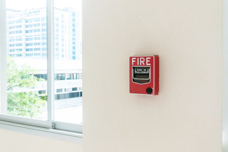 A red fire alarm mounted on a light wall beside a window, emphasizing safety and emergency preparedness in a modern indoor environment.の写真素材