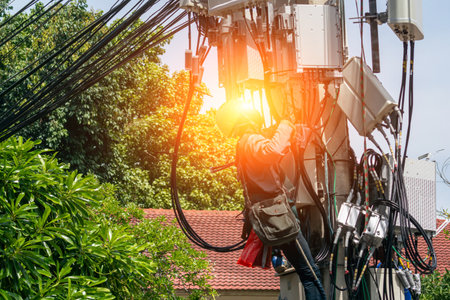 A technician working diligently on an electric pole, surrounded by wires and cables, ensuring safety and maintenance in a city environment under sunlight.の写真素材