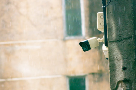 A close-up view of a surveillance camera mounted on a wall, captured during rain. This image...