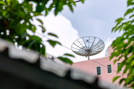 A satellite dish mounted on a rooftop under a cloudy sky. The image captures technology integrated into a residential setting, framed by greenery.の写真素材