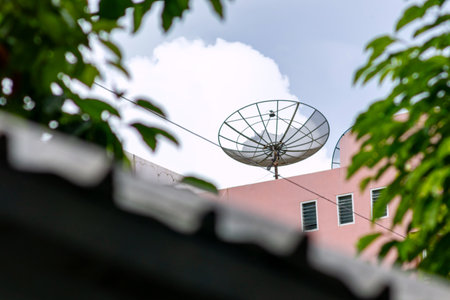 A close-up view of a satellite dish installed on a rooftop, surrounded by foliage and under a cloudy sky, showcasing modern communication technology.の写真素材