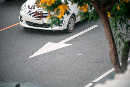 A white car is moving along a city road marked with a directional arrow. Lush foliage adds a touch...