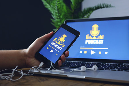 Wooden desk with a laptop computer with podcast website service in the screen. Hand holding smartphone with podcast app.の写真素材