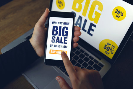 Closeup of smartphone with white screen and sign - Big Sale in female hand. In background blurred notebook. Woman using the gadget to surf Internet, online shopping, store app.の写真素材