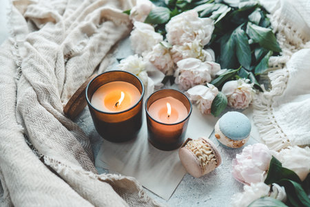 Two burning candles in a cozy home interior, white peonies and macarons.