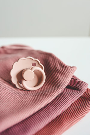 Stack of pink baby clothes and baby pacifier on white background.