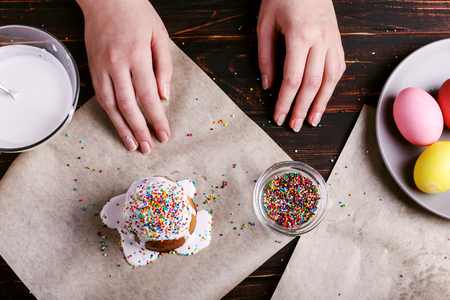 The girl prepares Easter baking, smears cake with icing and sprinkles with colored powder. Preparing for the holiday on the kitchen table, on a dark background.の写真素材