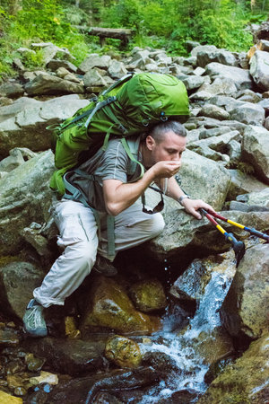 Hiker bending to take a drink from the stream in the mountain. Young hiker with green  rucksack and trekking pole drinking stream water with his hands,  in mountainの写真素材