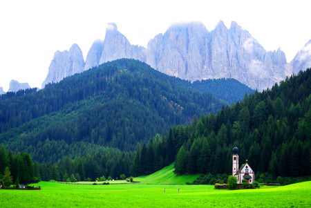 green meadow with small white church under the rocky mountains of the dolomites and the cloudy skyの写真素材