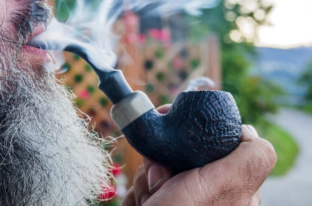 happy old man smokes the tobacco in his pipe with pleasure among sweet swirls and clouds of smoke