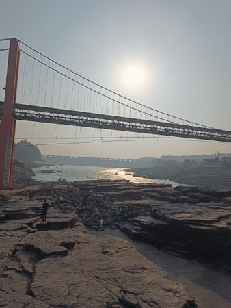 A golden sunrise over a suspension bridge and rocky riverbed. Sunlight reflects on the water as a lone person walks across the rocks, adding depth and perspective to this early morの写真素材