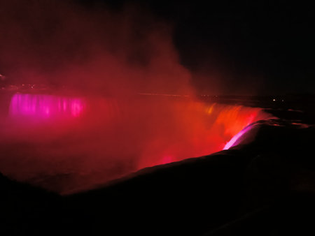 A dramatic night photo of Niagara Falls (Horseshoe Falls) illuminated in deep red light, with mist glowing against a dark black sky. The vibrant red glow creates a surreal, atmosphの写真素材
