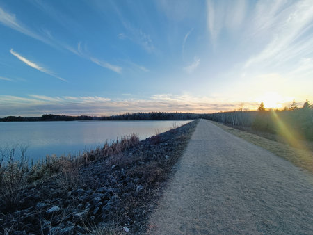 A scenic outdoor view featuring a dirt trail or unpaved road running alongside a body of water, with the setting sun glowing in the top right against a clear blue sky. The balancedの写真素材