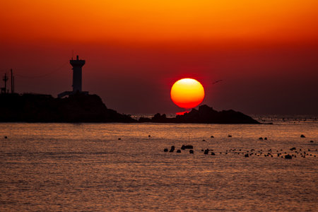 the sun rising between the lighthouse and the rocks of the seashoreの写真素材