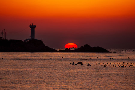 the sun rising between the lighthouse and the rocks of the seashoreの写真素材