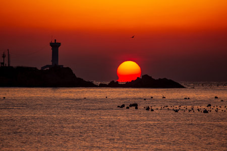 the sun rising between the lighthouse and the rocks of the seashoreの写真素材
