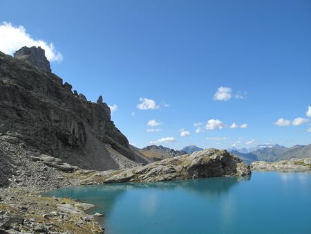 Gray mountain tops, cotton clouds and a deep blue sky are reflected in the sunny turquoise alpine...
