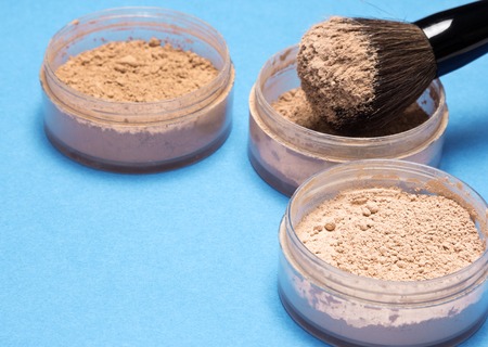 Jars filled with loose cosmetic powder and makeup brush on blue background. Focus on the contents of first jar. Copy spaceの写真素材