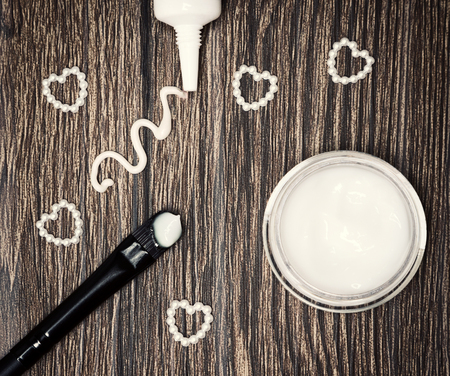 Favorite cosmetic products for skin around eyes. Eye cream squeezed out of tube, glass jar of light gel for eyelids and applying brush with sample and small hearts made of beads on wooden surfaceの写真素材