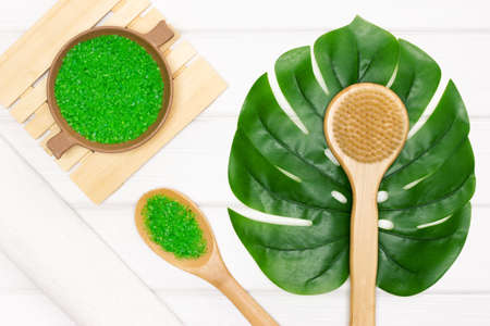 Body brush, sea salt, white towel with monstera green leaf on wooden boards. Top view, flat lay. Dry...