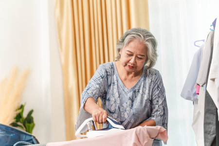 Portrait of a happy woman ironing clothes in living room at home, Cheerful Asian senior woman ironing on the tableの写真素材