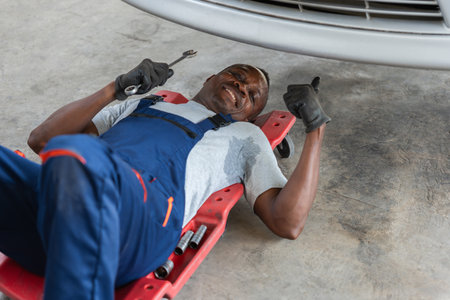 Happy Mechanic Under Car Giving Thumbs Up, Professional Auto Repairman Working in Garage, Sweaty Tradesman Fixing Vehicle on Creeperの写真素材