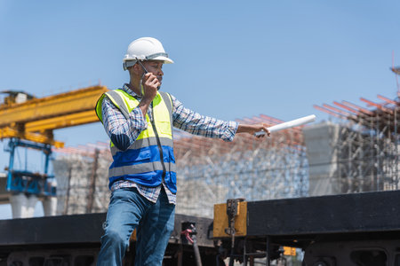 Construction Site Manager Communicating on Walkie-Talkie, Foreman with Plans and Radio at Building...