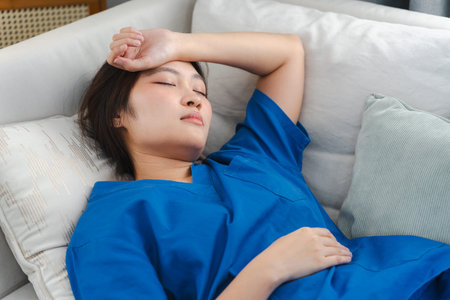 Exhausted young Asian nurse in blue scrubs resting on a sofa after a long shift, Tired medical...