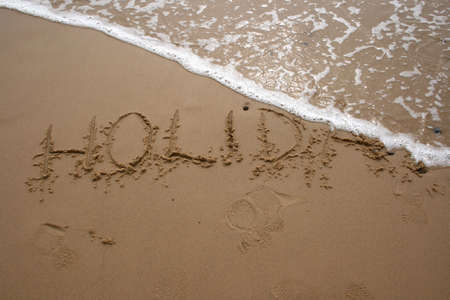 The word HOLIDAY written in sand next to the shore with a wave washing over の写真素材