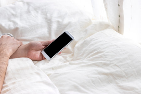 At hand of a man relaxing in bed and using a smartphoneの写真素材