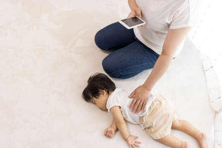 Mother using a smartphone and baby sleeping by the window (1 year old, Japanese, girl)の写真素材