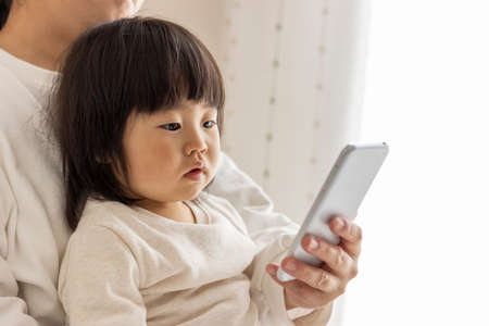 A baby looking at a smartphone (1 year and 8 months old, girl, Japanese) and a mother's hand showing it (30s, Japanese)の写真素材