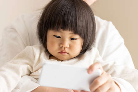 A baby looking at a smartphone (1 year and 8 months old, girl, Japanese) and a mother's hand showing it (30s, Japanese)の写真素材
