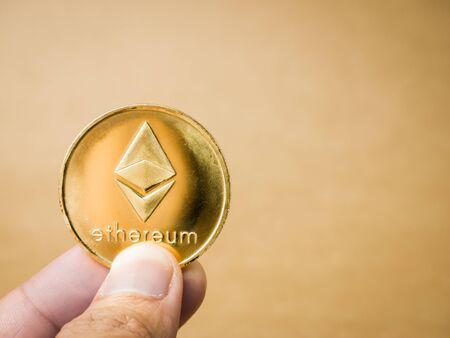 Hand is holding gold ethereum coin on a brown background. Business value ethereum coin are expensive.の写真素材