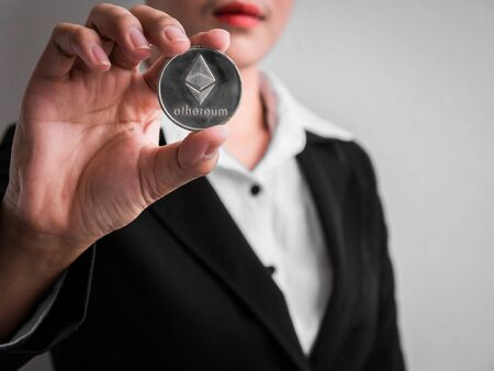 Businesswoman showing silver ethereum coin on the white wall is background. Investors woman holding...
