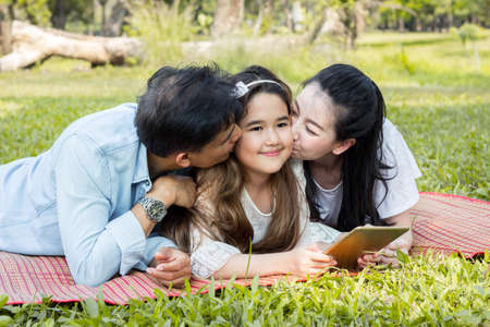 Parents and children are playing the tablet on the mat in the park. Father and mother are teaching by using the tablet. Girls are learning from a tablet that parents are teaching.の写真素材