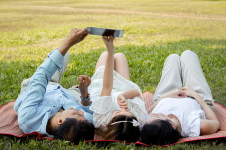 Asian family are using the tablet on the mat. Girls are learning from a tablet that parents are teaching. Parents and children are playing the tablet on the mat in the park.の写真素材