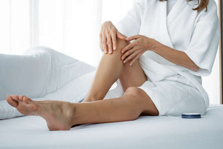 Beautiful young Caucasian woman applying body cream on her leg on white bed in the bedroom at home....