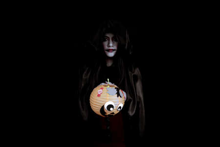 Young Asian woman costume in black witch with hand holding an orange Halloween pumpkin on black background with concept for Halloween fashion festival in studio. Teen Asian woman in cosplay Halloween.の写真素材
