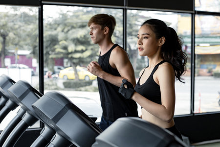 Young woman and man running side by side on modern electric treadmills at the gym. Pretty asian woman running on treadmill with fit young man on background. Young couple on electric treadmills.の写真素材