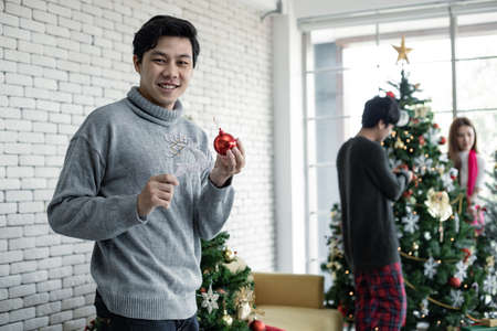 Young Asian man holds the toy to decorate the Christmas tree with smile on which has friends background. Celebration of new year at home. Merry Christmas and happy holidays.の写真素材