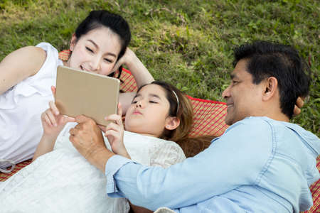 Asian family are using the tablet on the mat. Girls are learning from a tablet that parents are teaching. Parents and children are playing the tablet on the mat in the park.の写真素材