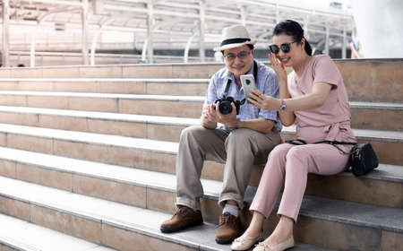 Senior Asian couples sit on the stairs while traveling and have fun video calling with someone. Senior couple travel concept.の写真素材