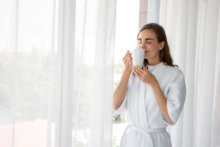 Beautiful Young Caucasian woman in pajamas holding a glass of white coffee ready to drink by the window in the bedroom at home after wake up in the morning. Woman standing drinking coffee by window.の写真素材