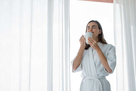 Beautiful Young Caucasian woman in pajamas holding a glass of white coffee ready to drink by the window in the bedroom at home after wake up in the morning. Woman standing drinking coffee by window.の写真素材