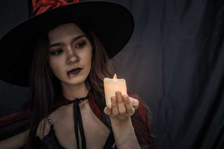 Young Asian woman in costume witch and hold candle on black cloth background of Halloween concept. Portrait of teen woman dressed up as witch for celebrate Halloween festival.の写真素材