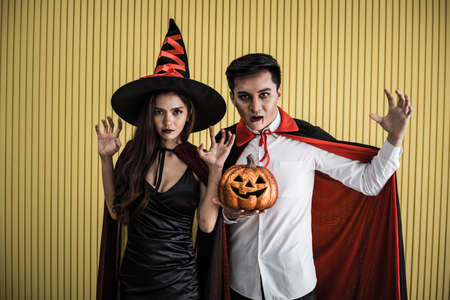 Halloween concept of young Asian woman in costume witch and Asian man in costume dracula on yellow background. Portrait teen couple dressed up as witches and dracula for celebrate  Halloween festival.の写真素材
