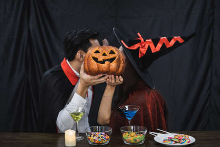 Young Asian couple in costume witch and dracula with celebrate Halloween party and kissed with a pumpkin covering the face. Couple in costume celebrate Halloween party black cloth background.の写真素材